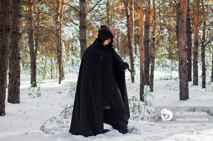 A warrior in black quilted clothes, in a black cloak with a hood on his head and a long sword in his hands against the background of winter forest and snow.