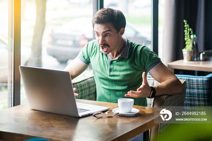 What do you want? Young angry businessman in green t-shirt sitting, working, looking and screaming at laptop screen on video call. business problem concept. indoor shot near big window at daytime.