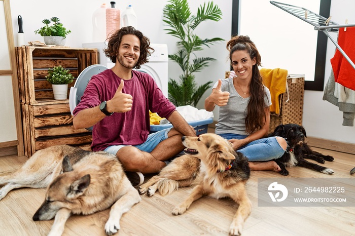 Young hispanic couple doing laundry with dogs doing happy thumbs up gesture with hand. approving expression looking at the camera showing success.