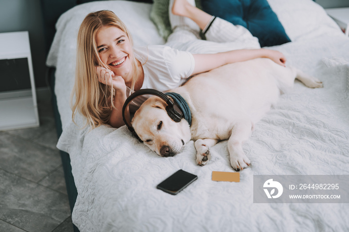 Delighted smiling beautiful woman lying in bed while having fun with her dog with headphones