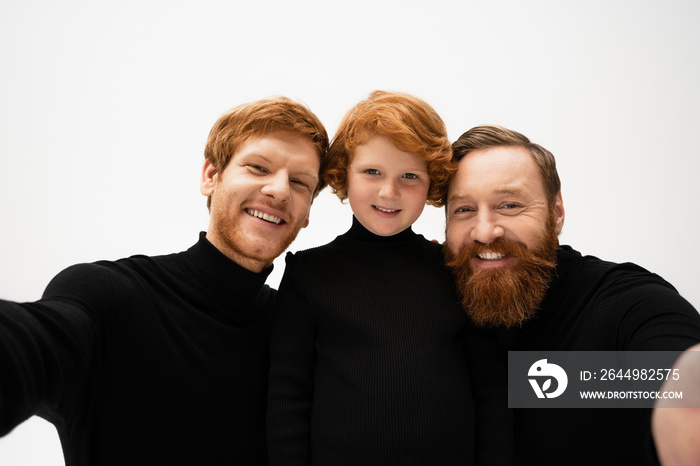 joyful bearded men in black pullovers smiling at camera near red haired boy isolated on grey.