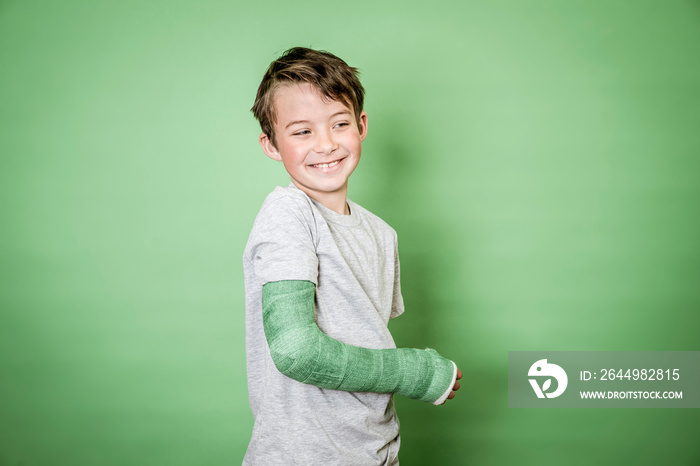 cool young schoolboy with broken arm and green plaster posing in front of green background in the studio