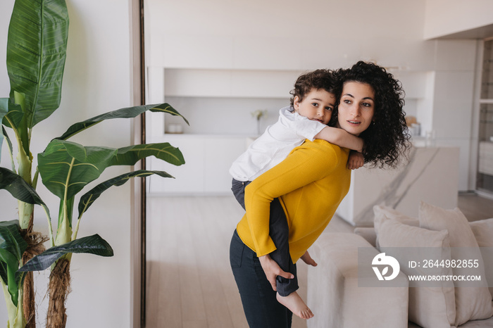 Young Spanish curly woman having fun with son, rolls him on back looking aside. Babysitter in yellow sweater and green pants entertains little cheerful child at home. Elder sister plays with brother.