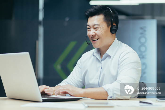 Learning online. Young handsome male Asian student studying remotely with laptop and headphones. Sitting at a desk in an office on campus.