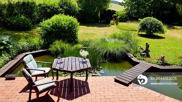 attraktiver naturbelassener Teich an rote Terrasse angrenzend mit Brücke und Tisch und Stühlen