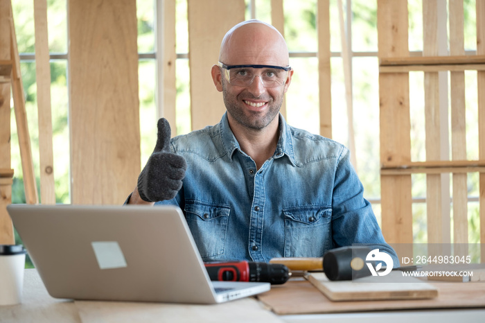 Confident senior craftsman carpenter wearing goggle and glove in the wood workplace. Looking forward and thumb up. There are laptop and many equipment on the table. DIY and handmade concept