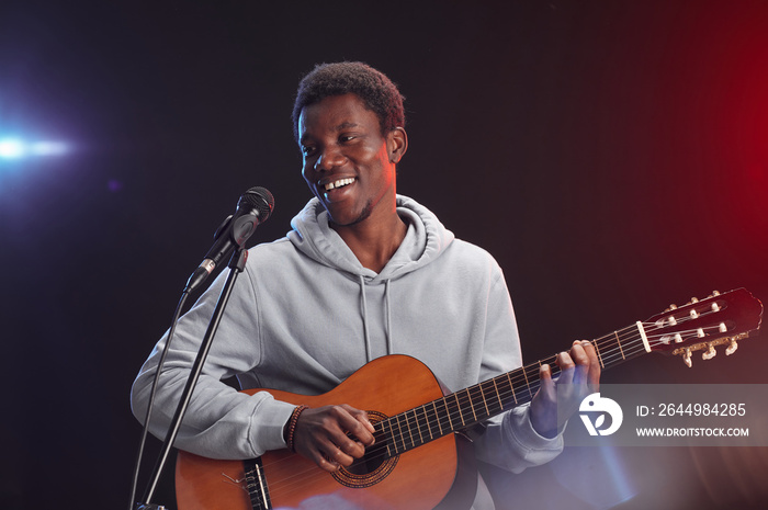 Waist up portrait of young African-American man playing guitar on stage and singing to microphone in lights, copy space