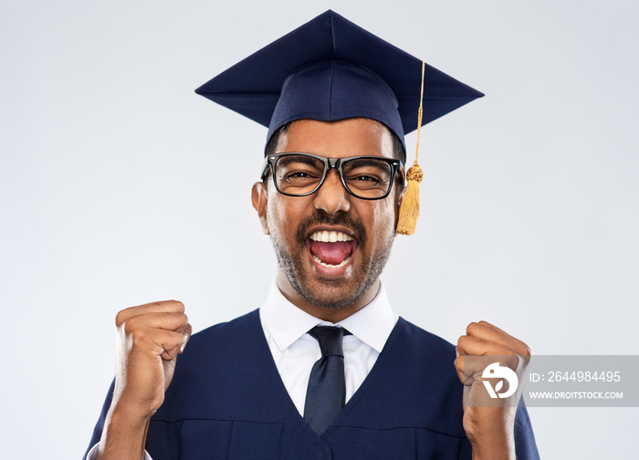 education, graduation and people concept - happy smiling indian male graduate student in mortar board and bachelor gown celebrating success over grey background