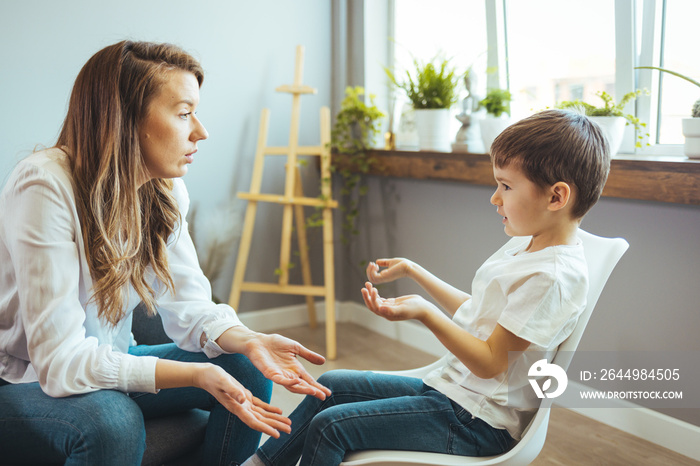 Family concept, mother and son relationship, happy young boy holding a teddy bear and talking to his mother. Smiling young woman talking to boy in white room