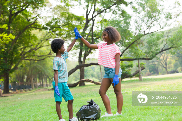 African American boy and girl wear gloves picking up plastic bottles into a black garbage bag outdoor in the park. Two children picking garbages outside. Volunteer and charity concept