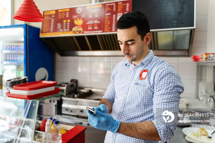 Portrait of caucasian man use phone app at fast food store - Young entrepreneur with protective gloves using mobile application to check online food orders - Mobile payment service real people concept