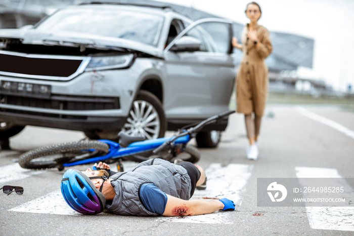 Road accident with injured cyclist lying on the pedestrian crossing near the broken bicycle and worried woman driver and car on the background