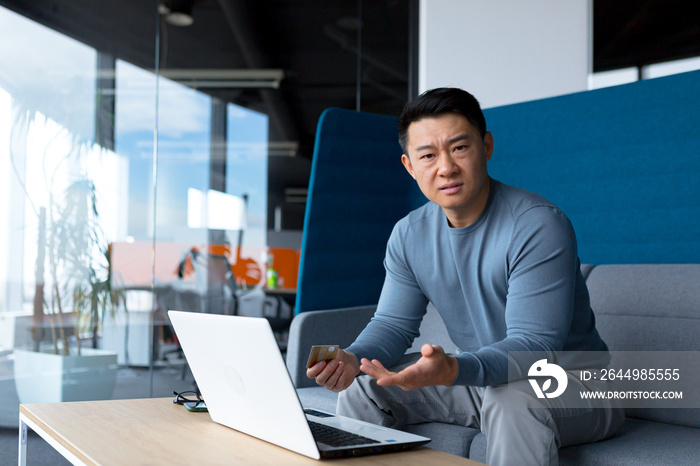 Annoyed and frustrated Asian man trying to make an online purchase sitting in the office at the computer, using a credit card