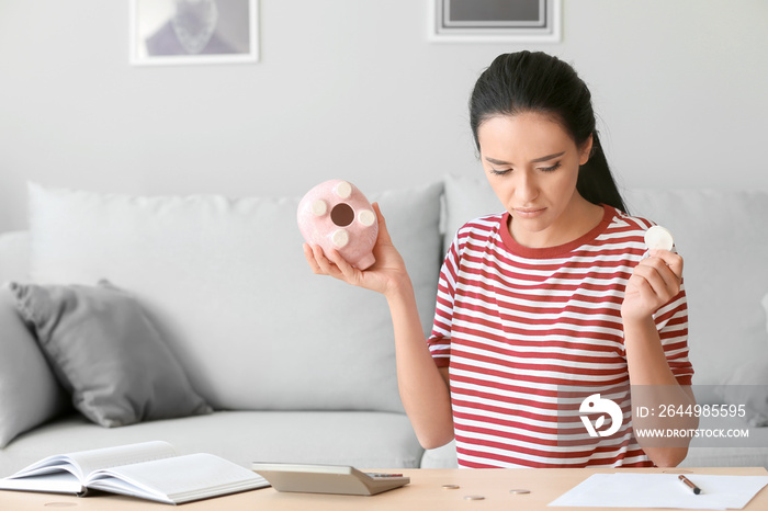 Upset young woman with empty piggy bank at home