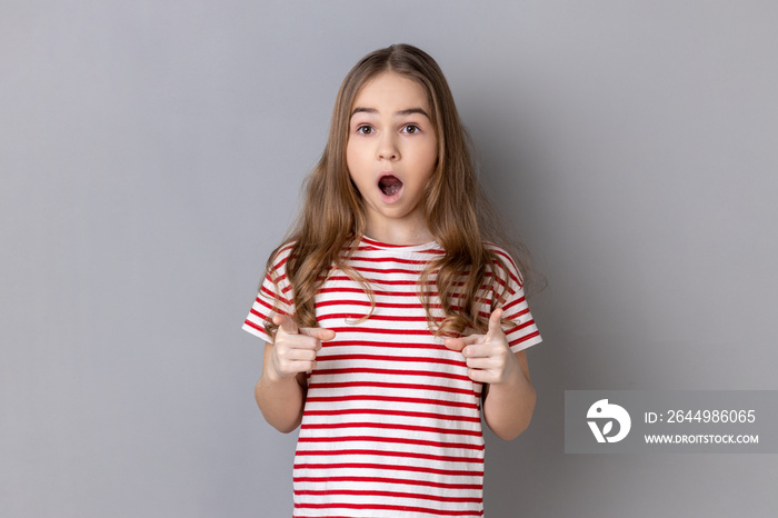 Portrait of astonished little girl wearing striped T-shirt pointing to camera with surprised shocked expression and open mouth, making choice. Indoor studio shot isolated on gray background.