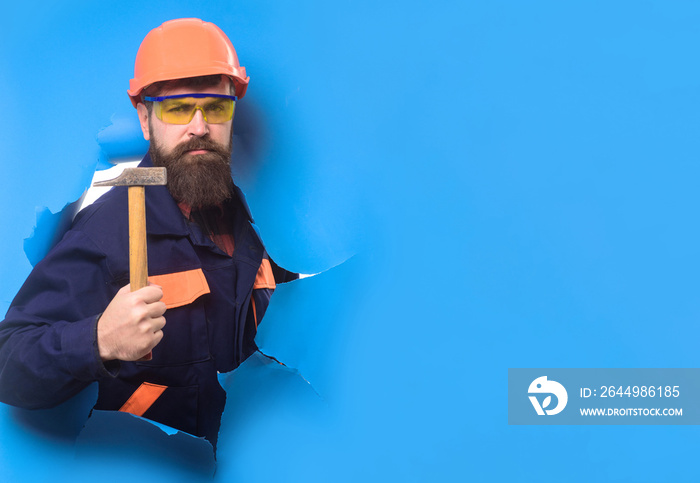 Bearded man in construction helmet makes repair. Builder in hard hat through hole in paper with hammer. Carpenter with hammer. Construction worker in hardhat. Home renovation. Repair home. Copy space.