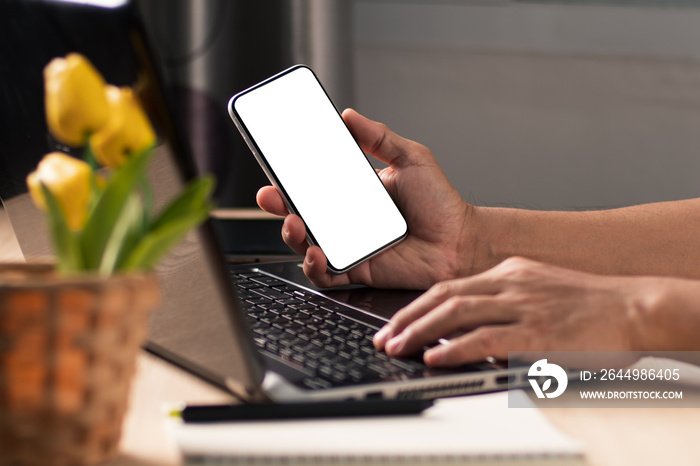 The businessman hand holding blank screen mobile phone during working on laptop computer at the home office.Mockup image of mobile phone.