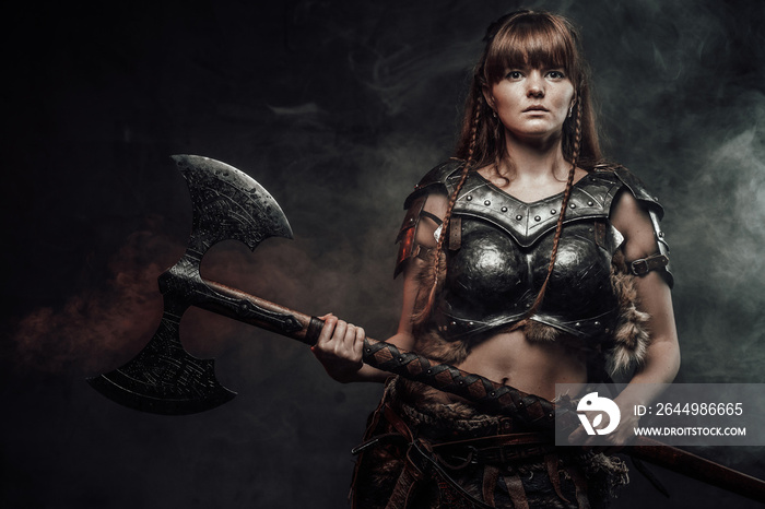 Atmospheric portrait of scandinavian amazon with brown hairs dressed in light armour holding two handed axe in dark background.