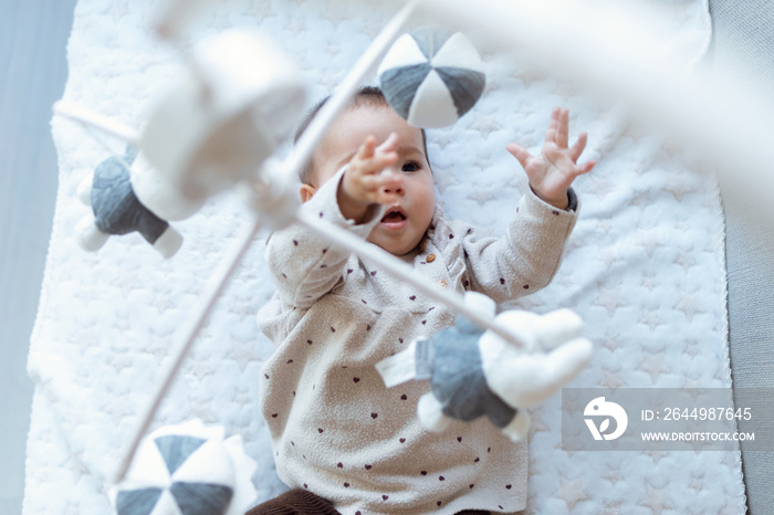 A cute baby girl is watching and playing with children’s musical mobile toy at home.