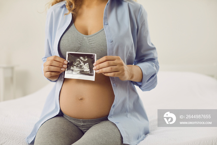 Young mother enjoying joys of motherhood. Happy pregnant lady sitting on bed and showing ultrasound ultrasonography echography scan of little human baby fetus growing and developing inside her belly