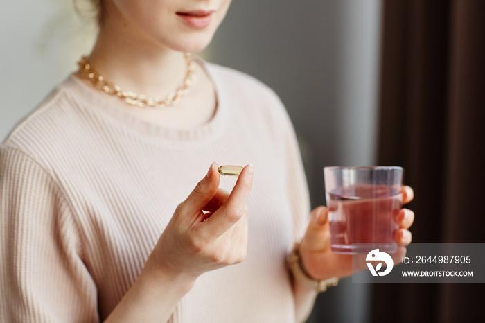 Minimal closeup of young woman holding vitamin capsule while taking supplements with glass of water at home