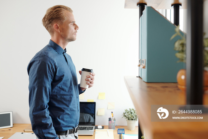 Young pensive businessman standing with cup of take out coffee and looking through office window