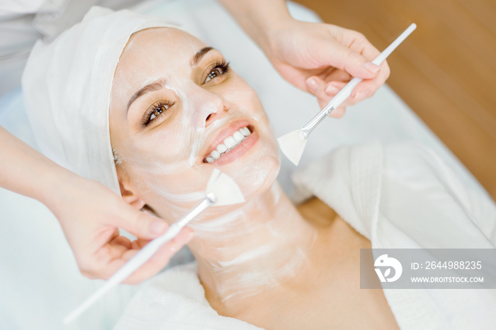 Cosmetology. The cosmetologist applies a cleansing face mask with two brushes. Smiling girl on the procedure for facial rejuvenation. Spa procedure.