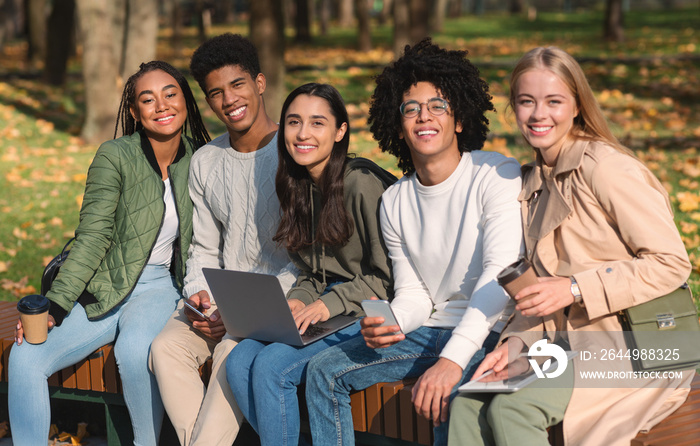 Cheerful teen friends sitting on bench with gadgets