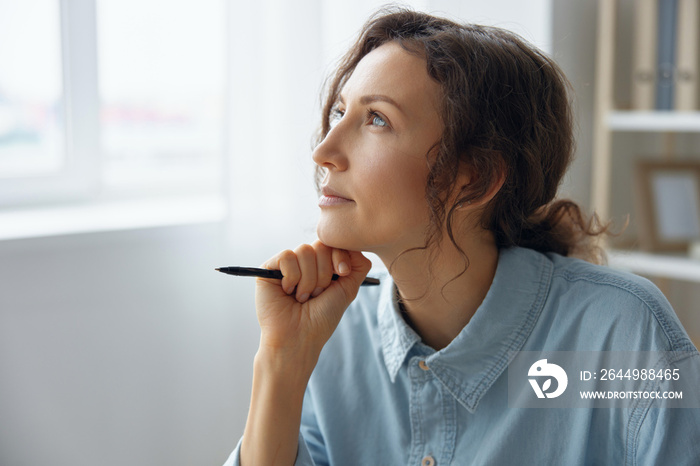 Portrait of pensive thoughtful dreaming curly adorable businesswoman looks aside pondering about right work decision can not choose reclines on fist with pen at office. Copy space for ad