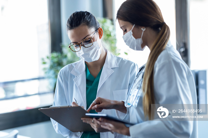 Two young women doctor wearing a hygienic facial mask talking with her colleague while working digital tablet in hospital.