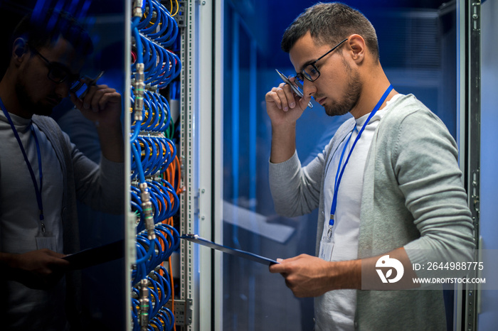 Side view  portrait of young man standing by server cabinet while working with supercomputer in data center and holding clipboard