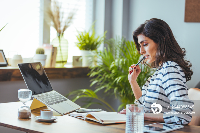 A shot of a young woman using a laptop and going through paperwork while working from home. A woman who uses a laptop while sitting at home. The concept of working from home