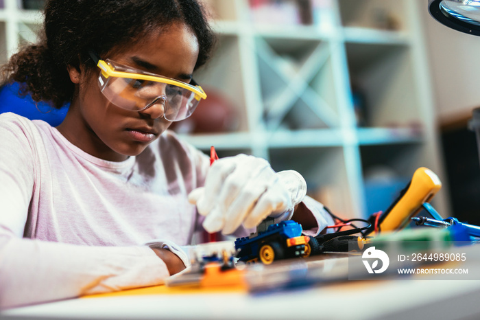 Smart Young African American Schoolgirl is Studying Electronics and Soldering Wires and Circuit Boards in Her Science Hobby Robotics Project. Girl is Working on a Robot in Her Room. Education Concept.