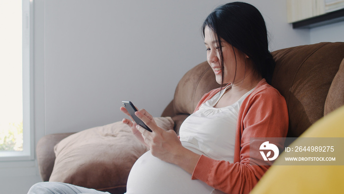 Young Asian Pregnant woman using mobile phone search pregnancy information. Mom feeling happy smiling positive and peaceful while take care her child lying on sofa in living room at home concept.