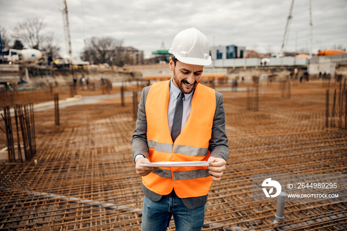 A happy site manager is visiting reconstruction area. A man is standing on building foundation and looking at the blueprints.