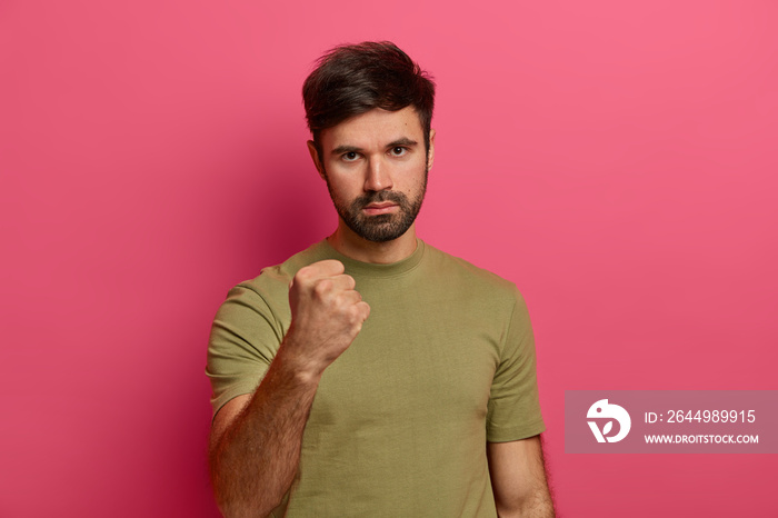 Angry strict man with thick beard, being furious, expresses warning and threat, shows his power or resoluteness, wears casual t shirt, isolated on pink background, looks with serious expression.