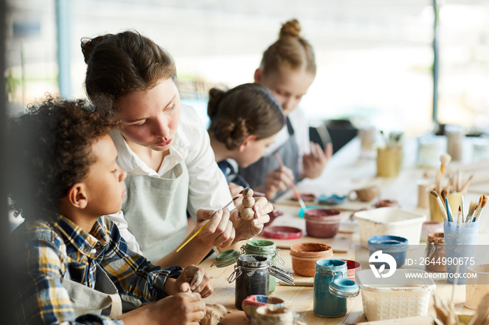 Young teacher showing one of pupils clay item and giving advice about painting it