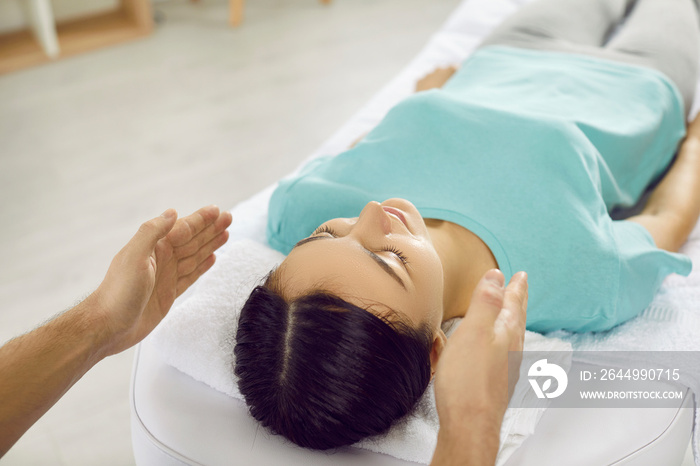 Woman undergoes alternative therapy during reiki session in modern massage parlor. Therapist holds his hands near head of beautiful young woman to transfer energy during Reiki healing session.