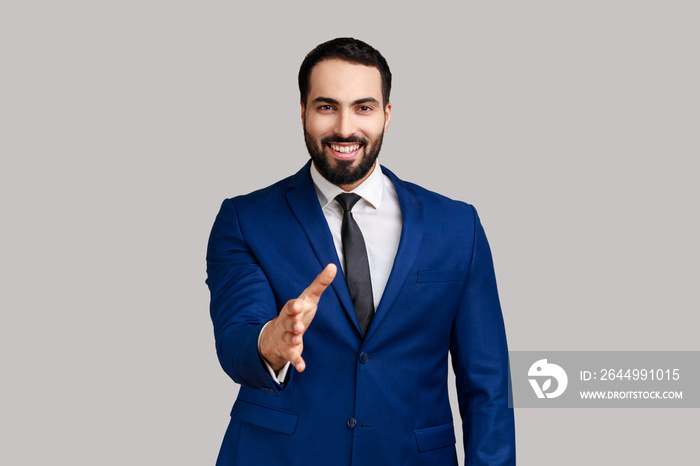 Positive bearded man giving hand to handshake and smiling friendly, consultant or HR manager offering job, partnership, wearing official style suit. Indoor studio shot isolated on gray background.