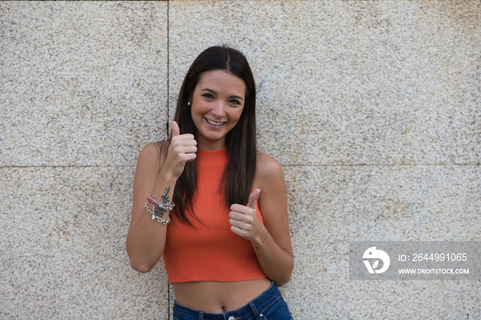 Beautiful brunette girl with long hair is happy and shows both hands with thumbs up because everything is correct and ok, isolated on beige background and orange top.