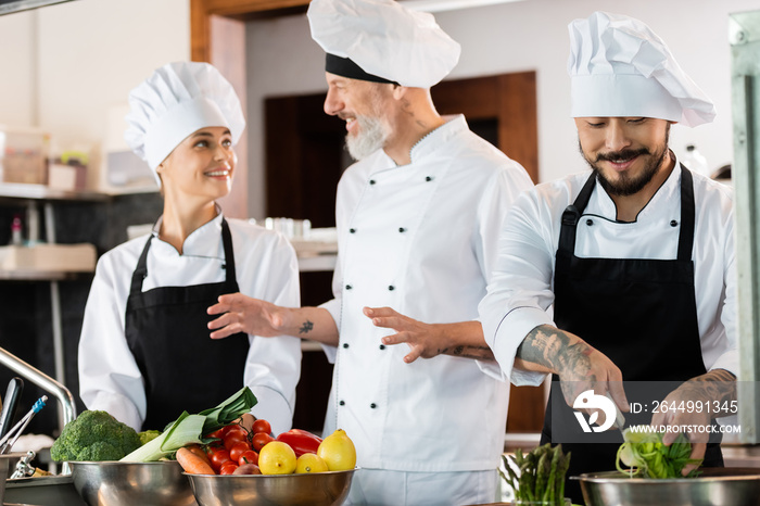 Smiling asian chef holding sliced leek while colleagues talking in kitchen