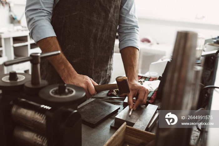 Important stage in the work. Male goldsmith wearing apron making a new product at a workbench using hammer