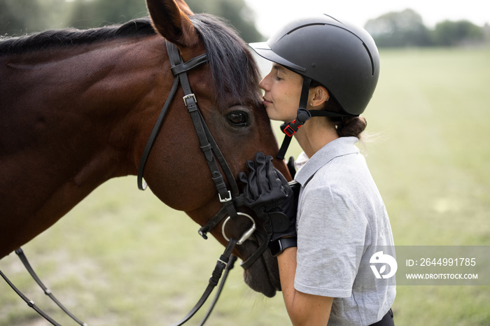 Female horseman hugging head of her brown Thoroughbred horse. Blurred image of green meadow in countryside. Concept of animal care. Rural rest and leisure. Idea of green tourism. Young european woman