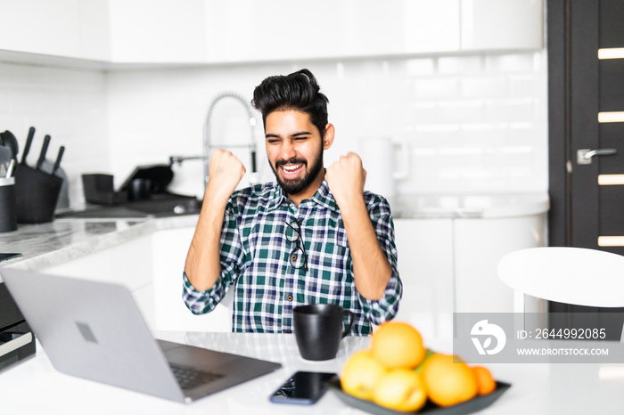 Young indian bearded man celebrating success while working on laptop in kitchen