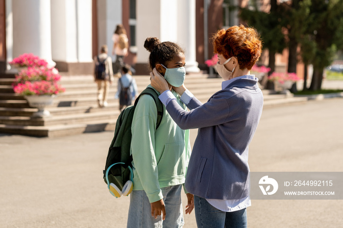 Young foster mother seeing mixed-race daughter off for school while putting protective mask on her face