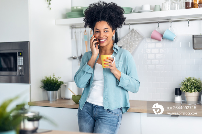 Smiling young woman talking with her mobile phone while drinking a cup of coffee in the kitchen at home.