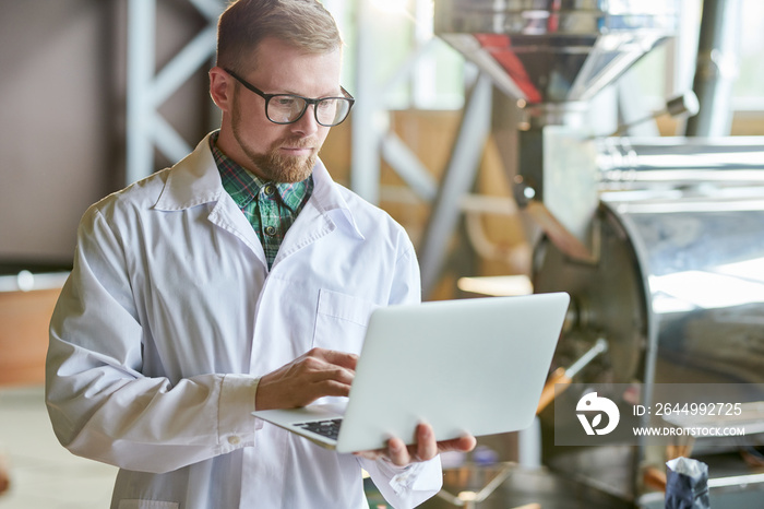 Waist up portrait of modern young man wearing lab coat using laptop while working in coffee production manufactory, copy space