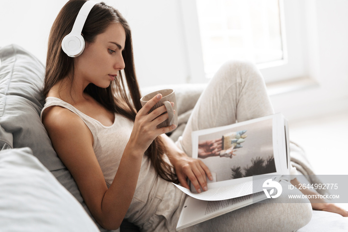 Young woman sitting on sofa indoors at home listening music with headphones drinking tea reading magazine.