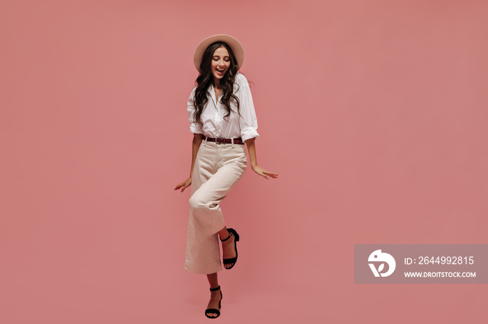 Cute curly black haired woman in white long sleeve shirt, light pants with brown belt, dark heels and modern hat looking up..