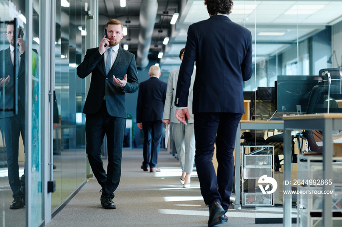 Young businessman in suit talking on mobile phone while walking along office corridor among business people